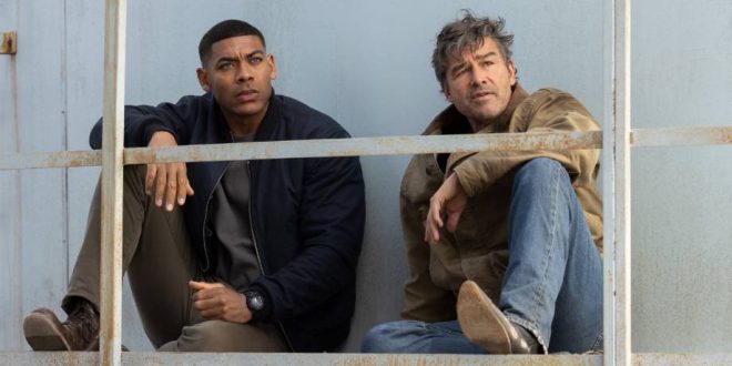 Two men sit on a metal platform against a weathered, pale blue wall, looking thoughtfully into the distance. One wears a dark jacket and watch, the other a tan coat and jeans.